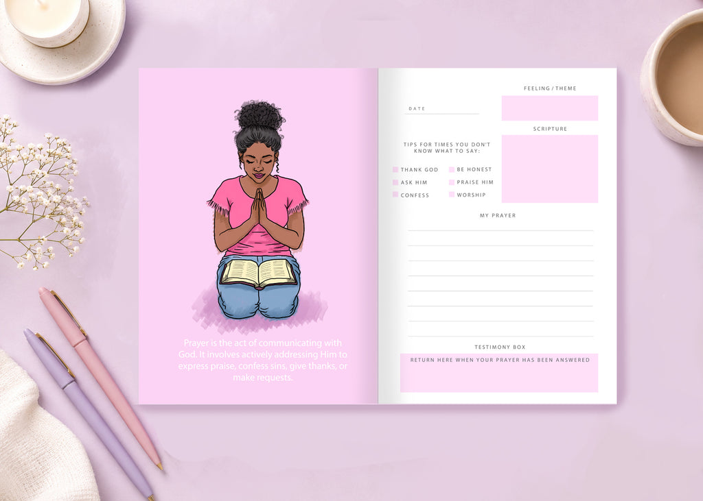 An open planner or journal spread on a pastel pink surface, showing a pink cover with an illustrated woman kneeling and reading, plus interior pages with prayer and reflection boxes. Decorative pens, flowers, and cups are arranged around it.