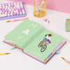 A pastel green open notebook or planner spread on a white desk, surrounded by pink and lavender stationery items including pens, clips, a pencil case, and part of a keyboard against a pink and white background.